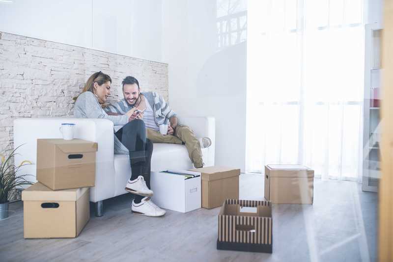 A couple look on a smartphone with cardboard moving boxes in their living room